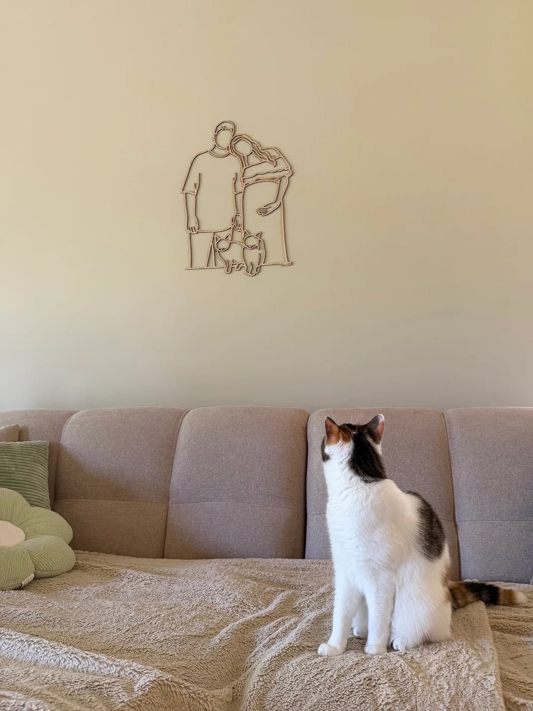 Katze auf dem Sofa blickt hinauf zum Linienporträt aus Holz ihrer Besitzer mit ihren zwei Katzen