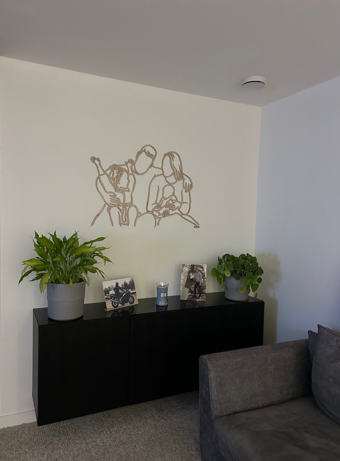 Linienporträt aus Holz einer Familie über einem schwarzen Sideboard, daneben Fotos von Papa